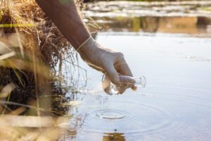 A person wears a rubber glove and fills water from a river into a beaker.