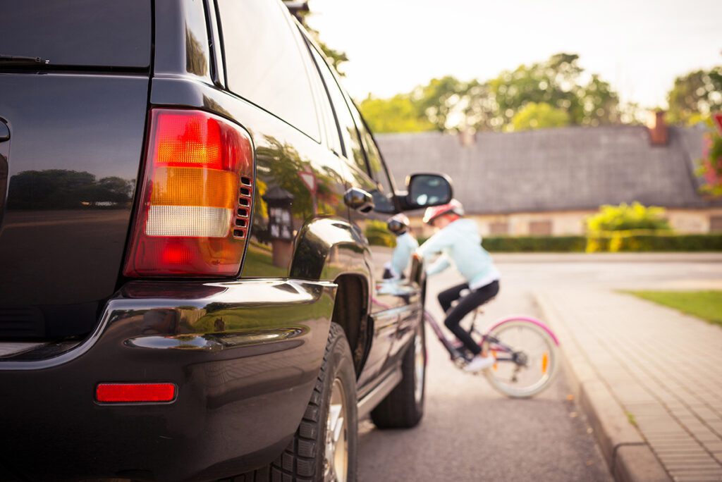 A child on a bike wearing a helmet rides out into the street in front of a black SUV.